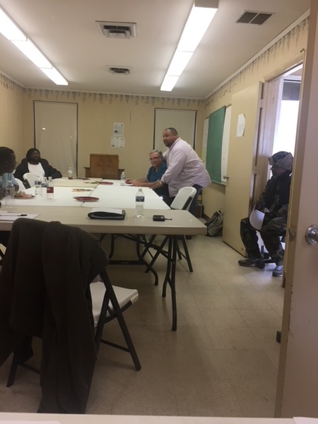 People sitting at a table listening to man leaning against the table.